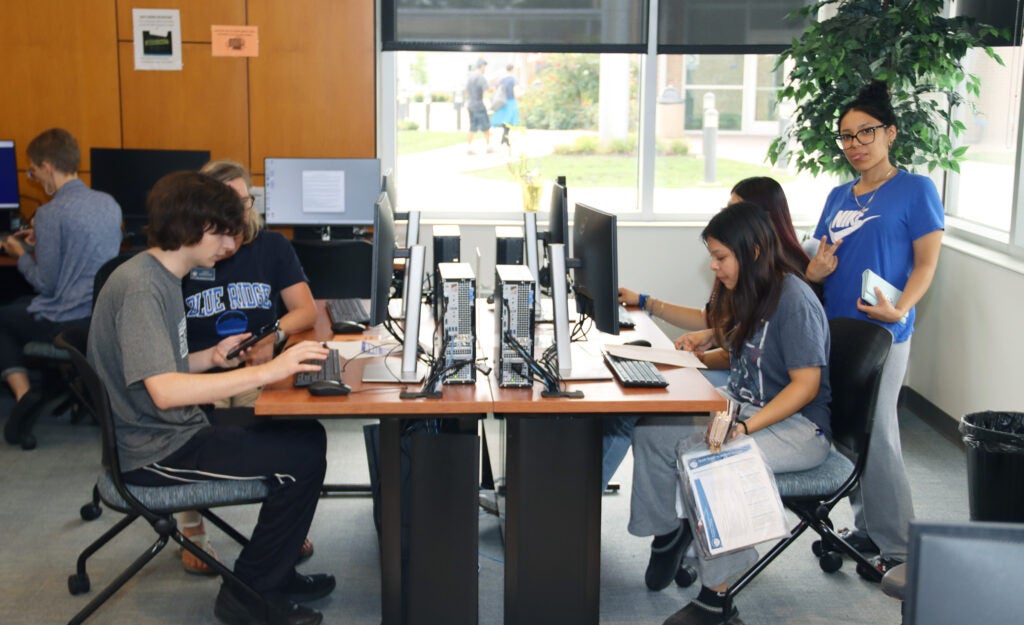 A row of computers set up on tables in front of a large window. A young man and a young woman are seated at computers with another young woman standing beside