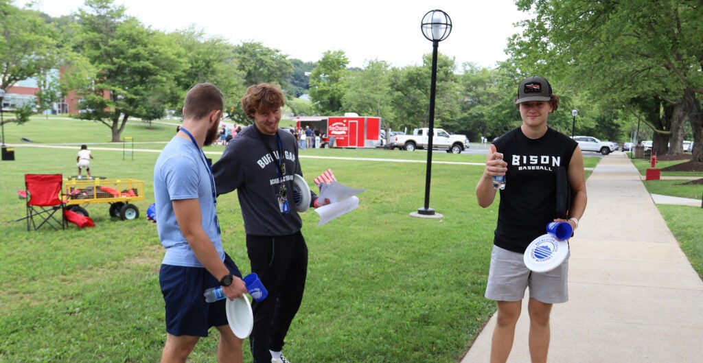 Three young men with frisbees talking on the sidewalk in front of a picnic table on the quad. An ice cream food truck is seen in the background.