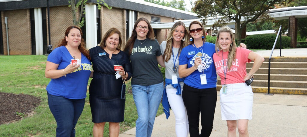A group of smiling women with their arms around each other in front of a campus building