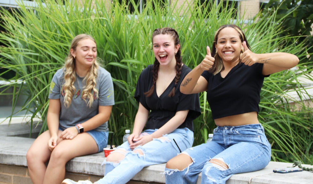 Three smiling young women sitting on a wall. One is giving two thumbs up.