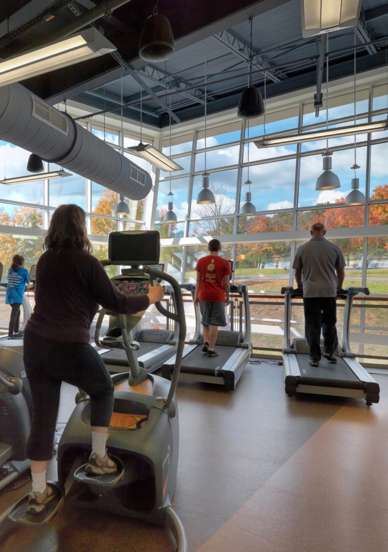 a woman on an elliptical trainer and two men on treadmills, facing a large window with brilliant fall foliage showing outside