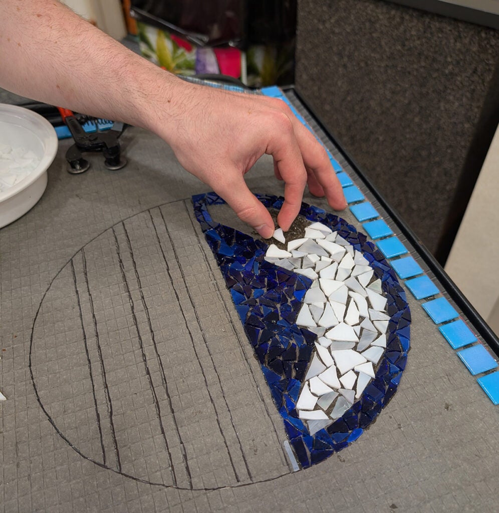 Part of the mosaic in progress, a hand placing white tile pieces in the BRCC logo