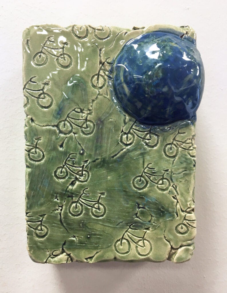 Ceramic sculpture of green rectangle with bicycles stamped in it and a blue-green globe at the top right corner, entitled "The Wheels on the Bike Go Round and Round" 2024 by Jessica Martinkosky